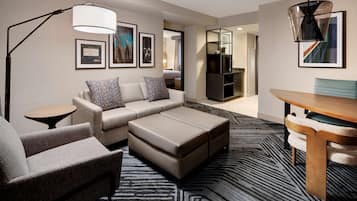 Una habitación de hotel moderna con sofá, reposapiés y una mesa de comedor. Hay una lámpara, una obra de arte enmarcada en la pared y un televisor al fondo.