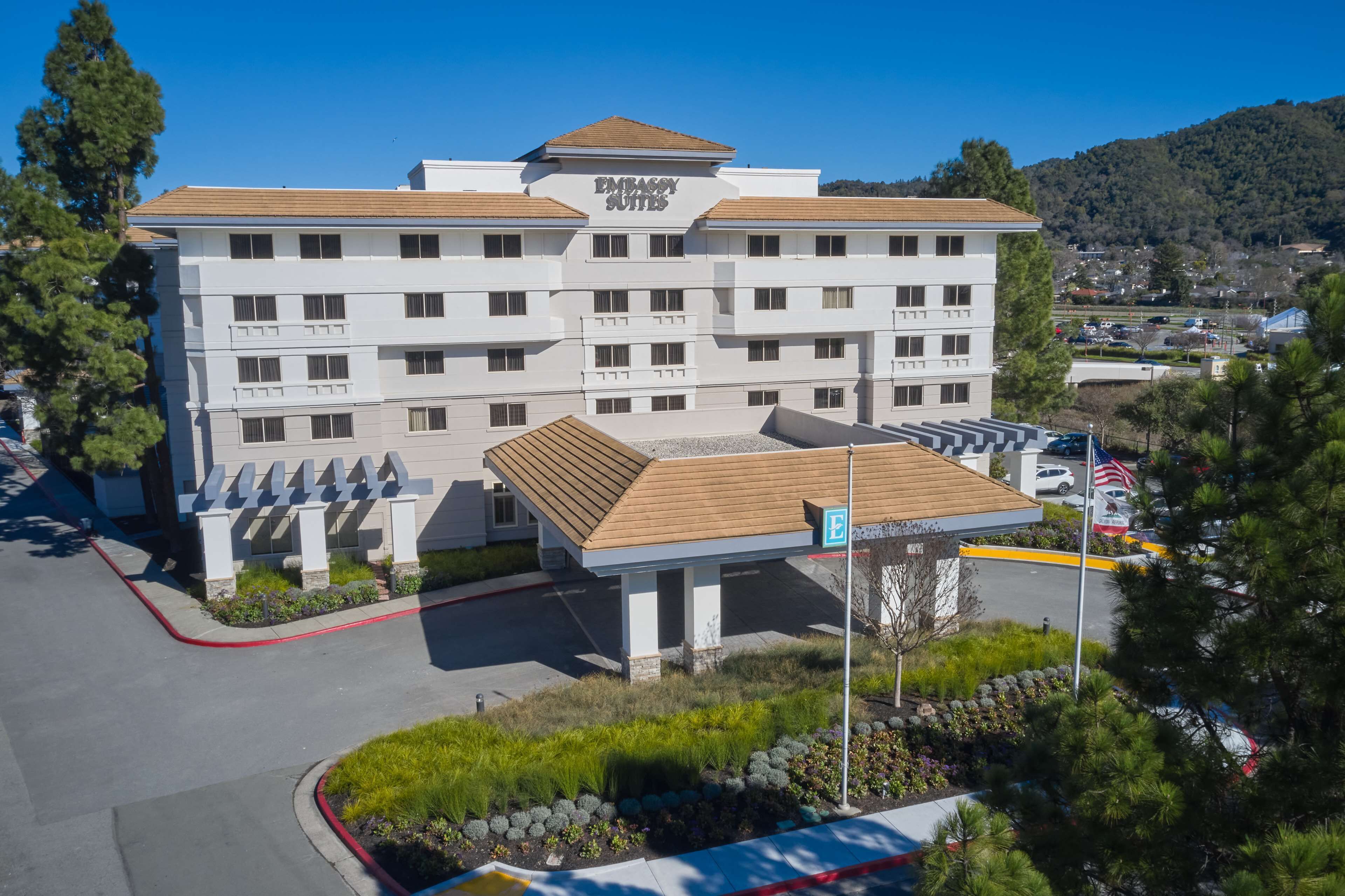 A multi-story building with a sign reading "Embassy Suites." It features a parking lot with a gas station, surrounded by greenery and a hilly landscape in the background.