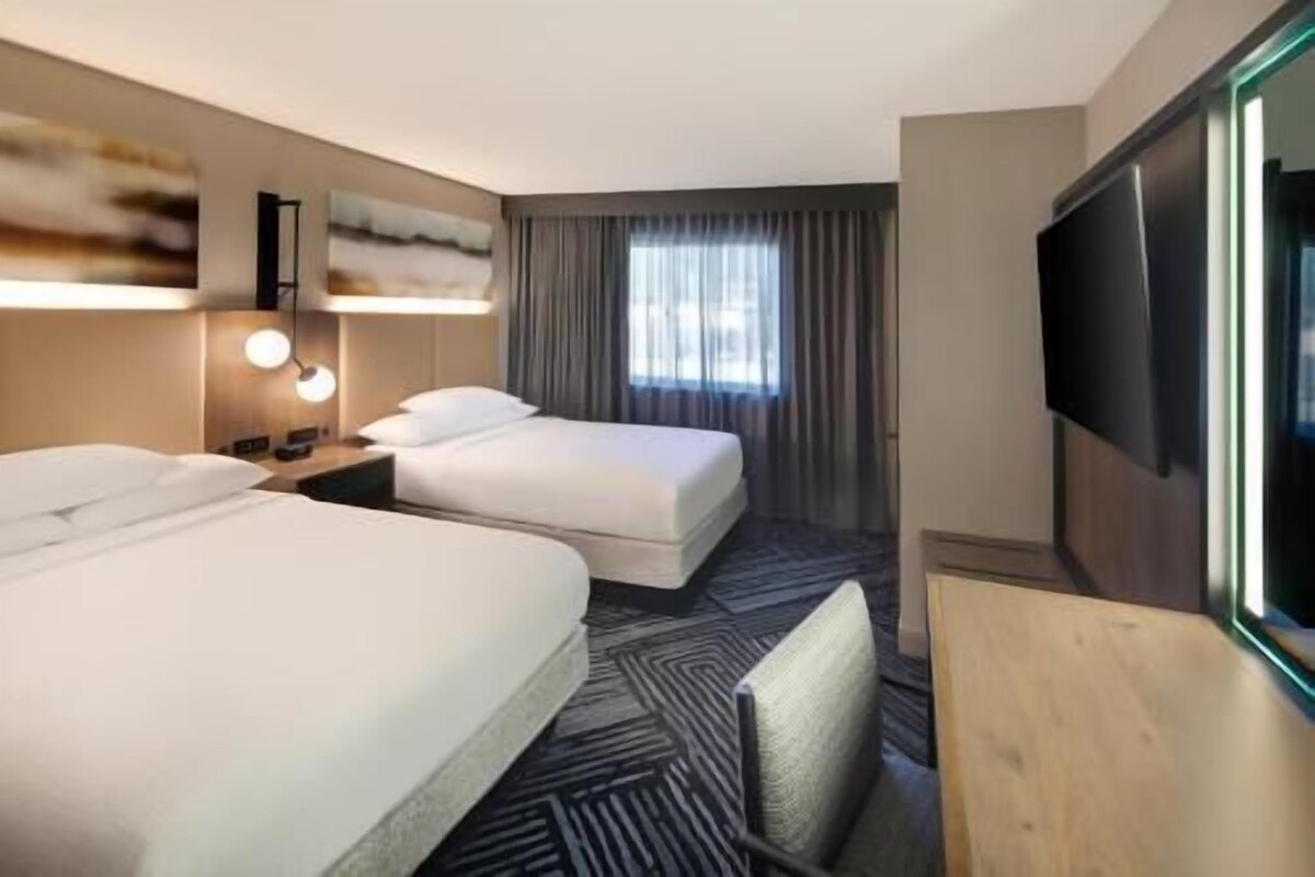 Ein Hotelzimmer mit zwei Betten, einem Fernseher, einem Schreibtisch und einem Fenster mit Vorhängen.