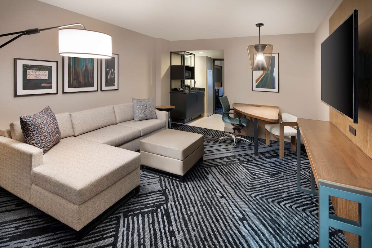 A modern hotel room with a sectional sofa, a desk, a flat-screen TV, and a bar area.