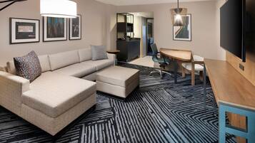 A modern hotel room with a sectional sofa, a desk, a flat-screen TV, and a bar area.