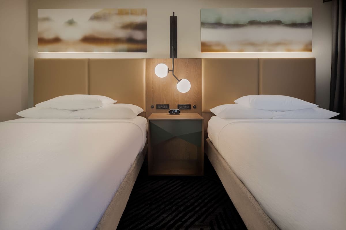 A hotel room with two beds, a nightstand with a lamp, and artwork on the wall.