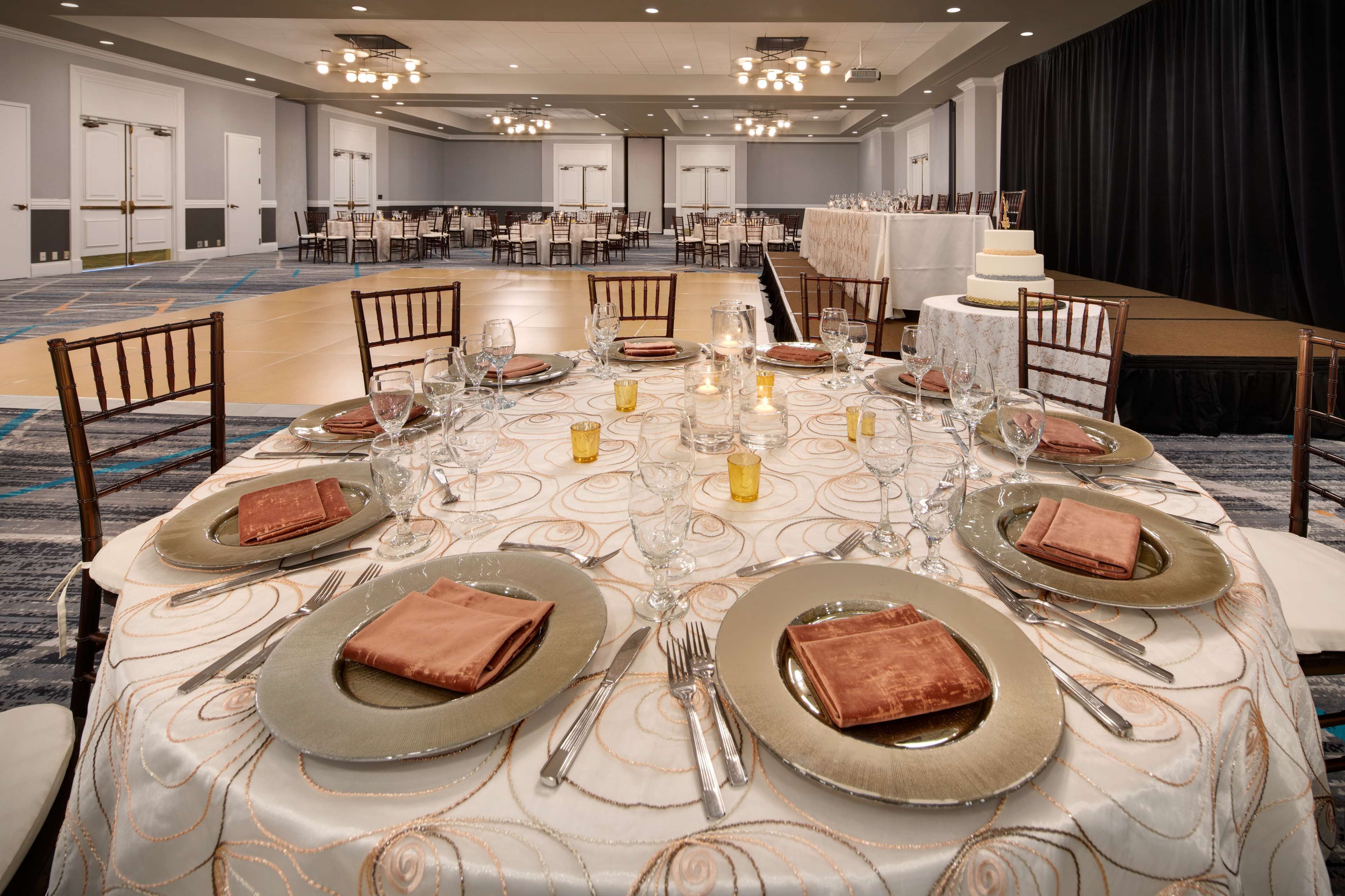 A banquet hall with round tables set for a formal event, featuring gold and brown napkins, clear glassware, and candles.