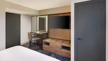 A hotel room with a bed, a desk with a chair, a large mirror, and a wardrobe.