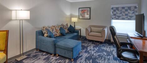 A hotel room with a sectional sofa, a chair, a desk, and a lamp.