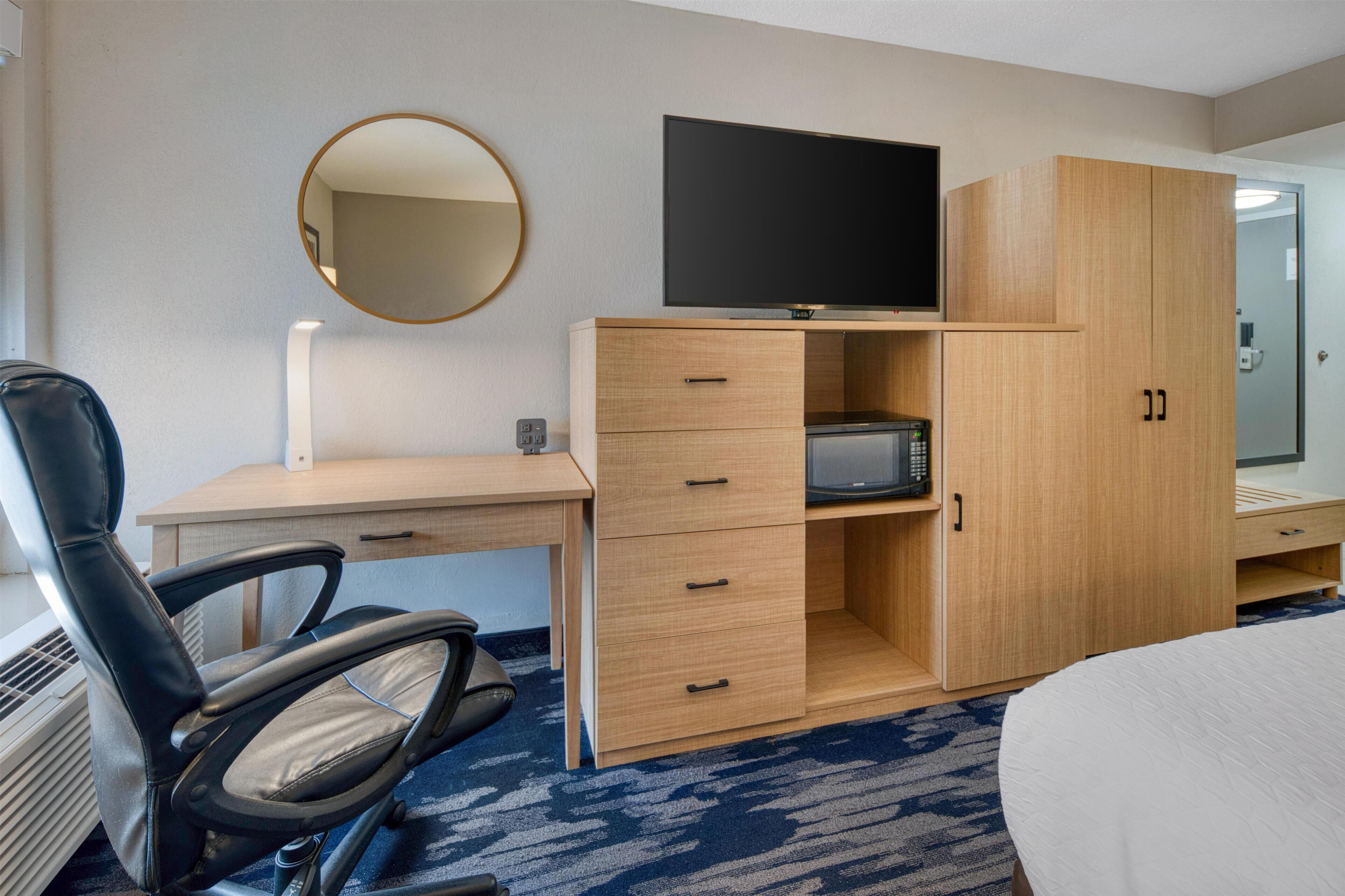 Una habitación de hotel con cama, escritorio y silla, televisor, armario y espejo.