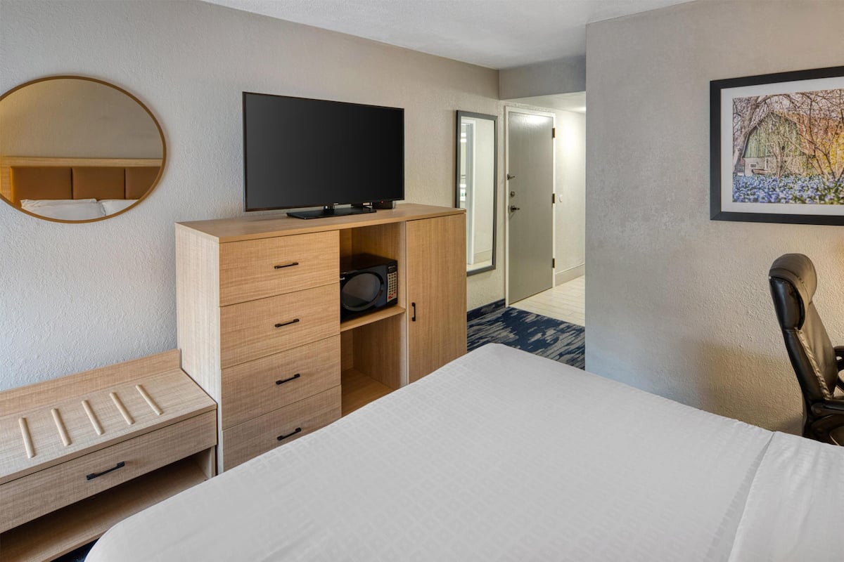 Ein Hotelzimmer mit einem Bett, einem Fernseher, einer Holzkommode, einer Mikrowelle, einem Spiegel, einer Tür und einem gerahmten Bild an der Wand.