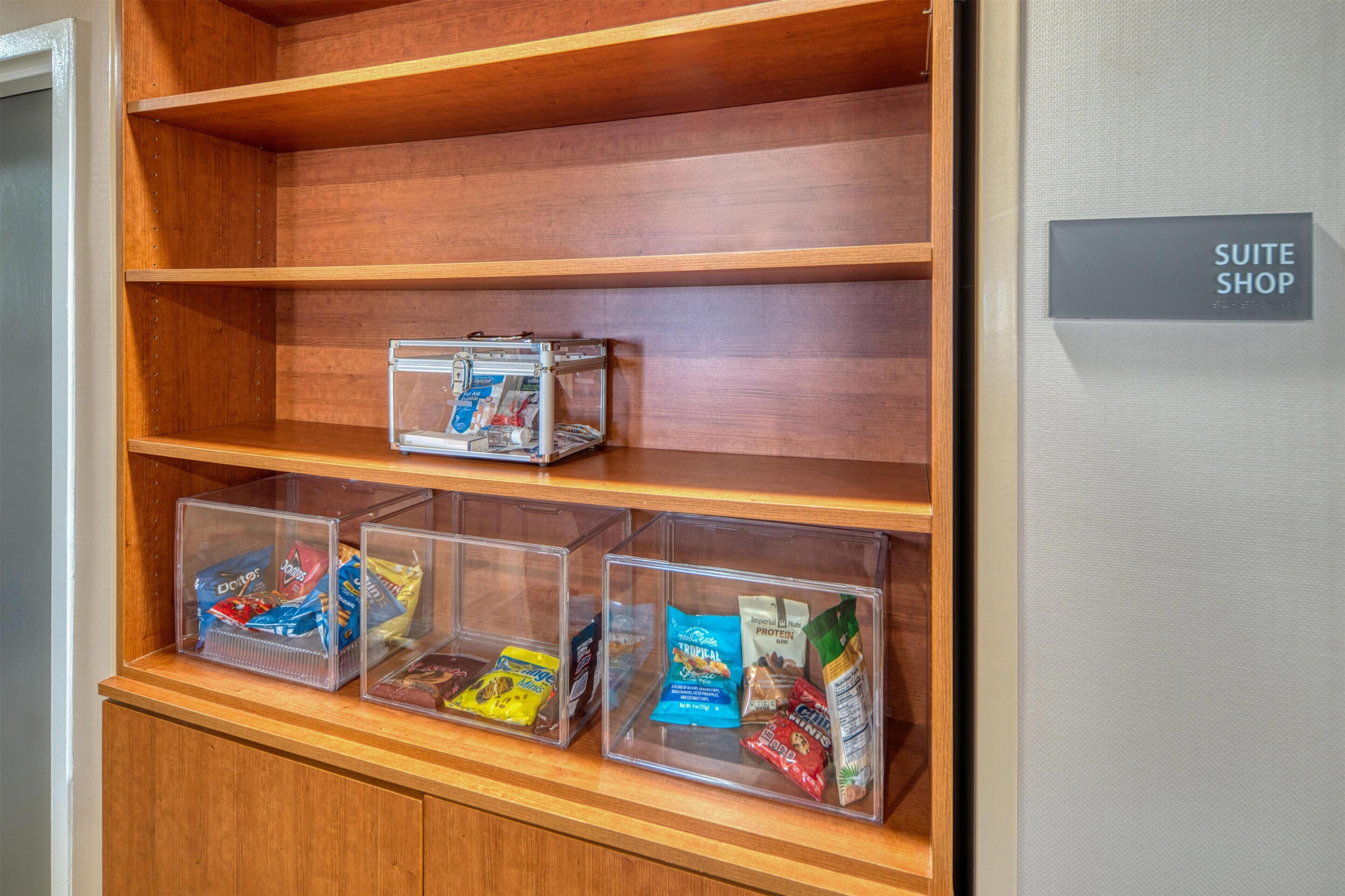 Een houten plank met doorzichtige opbergdozen vol snacks en een bordje met de tekst 'SUITE SHOP'.