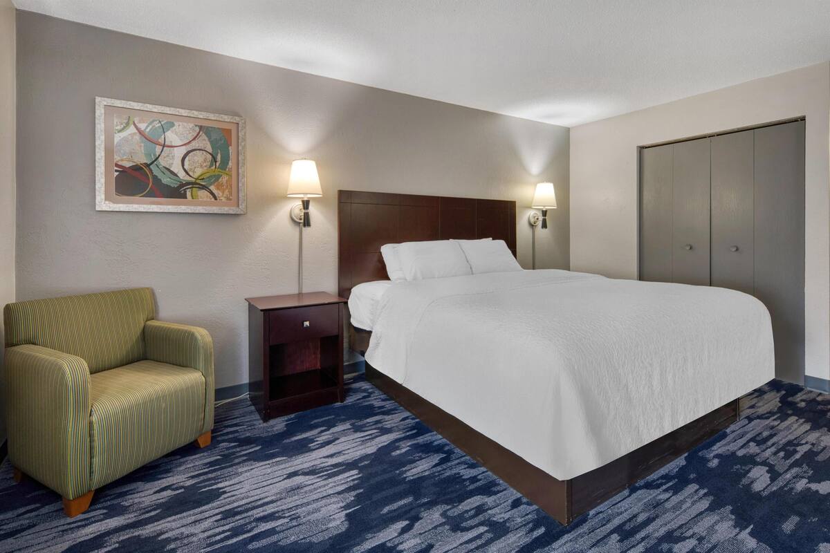 Ein Hotelzimmer mit Bett, Stuhl, Nachttisch, Wandbild und Kleiderschrank.