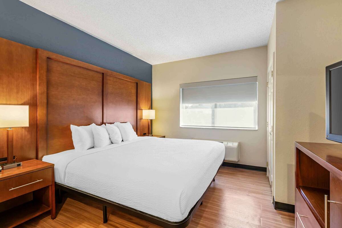 A hotel room with a large bed, wooden headboard, bedside lamps, a TV, and a window with blinds.
