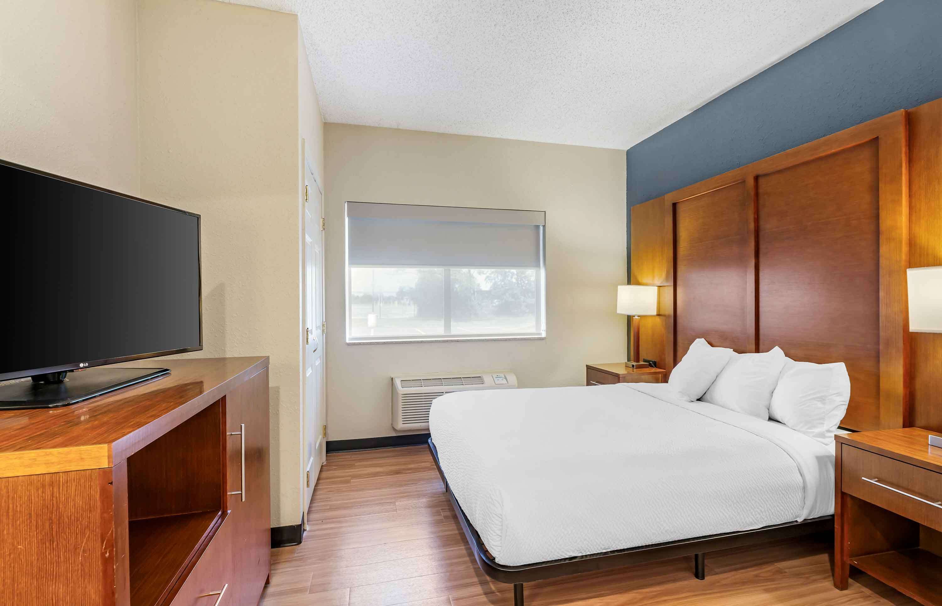 A hotel room with a bed, a TV on a wooden cabinet, a window with blinds, a bedside table with a lamp, and a wall-mounted heater.