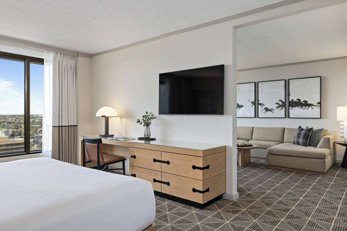 A hotel room with a bed, a desk with drawers, a TV, a sofa, and framed artwork on the wall.