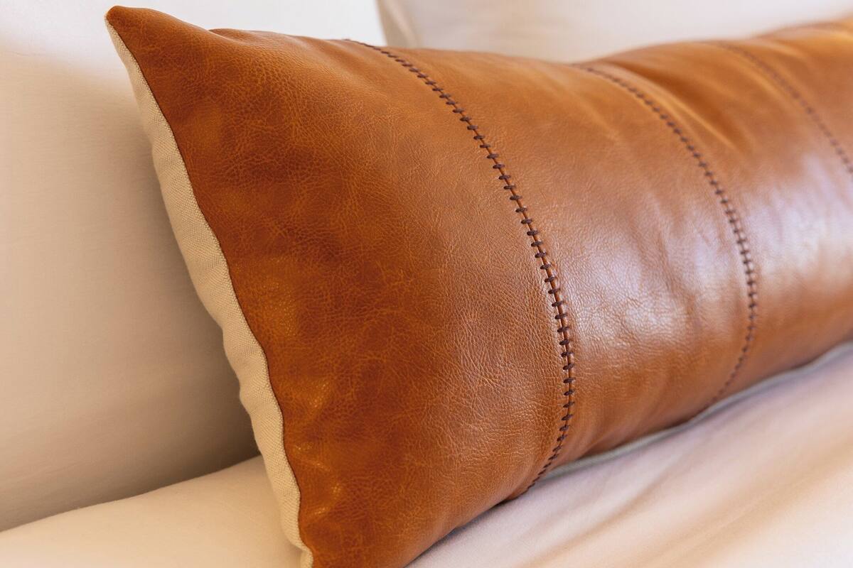 A close-up of a brown leather pillow with visible stitching on a white upholstered headboard.