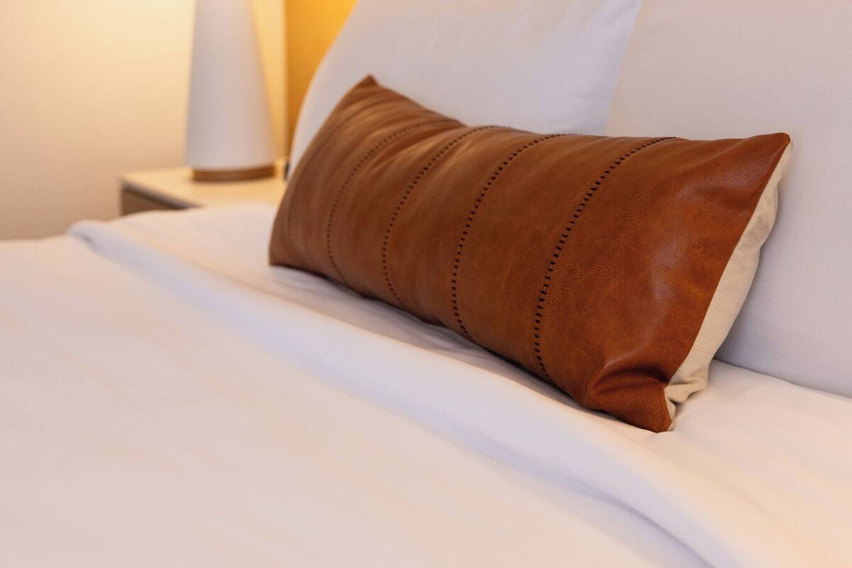 A neatly made bed with white linens and a brown leather pillow, a white lamp on a bedside table, and a textured wall behind the bed.
