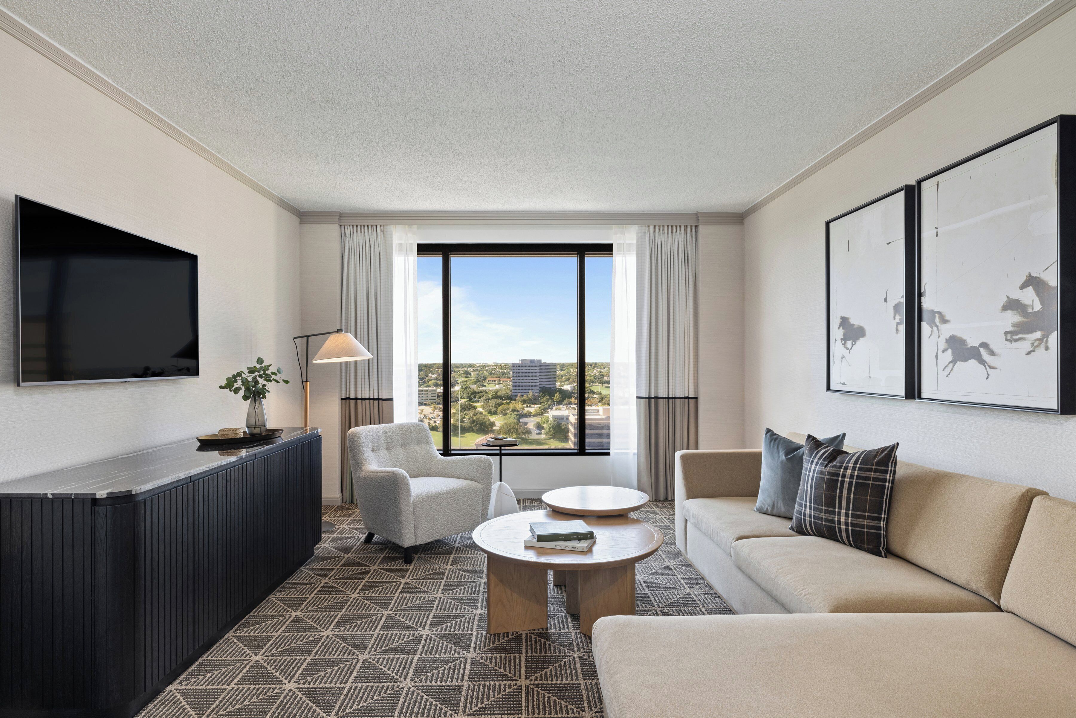 A modern hotel room with a large window, a flat-screen TV, a sofa, a coffee table, and a side table with a lamp.