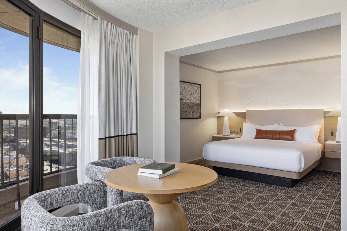 A hotel room with a bed, a round table, two armchairs, a large window with curtains, and a city view.