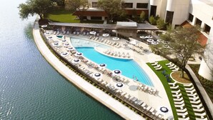 A resort pool area with multiple sun loungers and umbrellas, surrounded by a landscaped garden and a building in the background. - Omni Las Colinas Hotel (Irving)