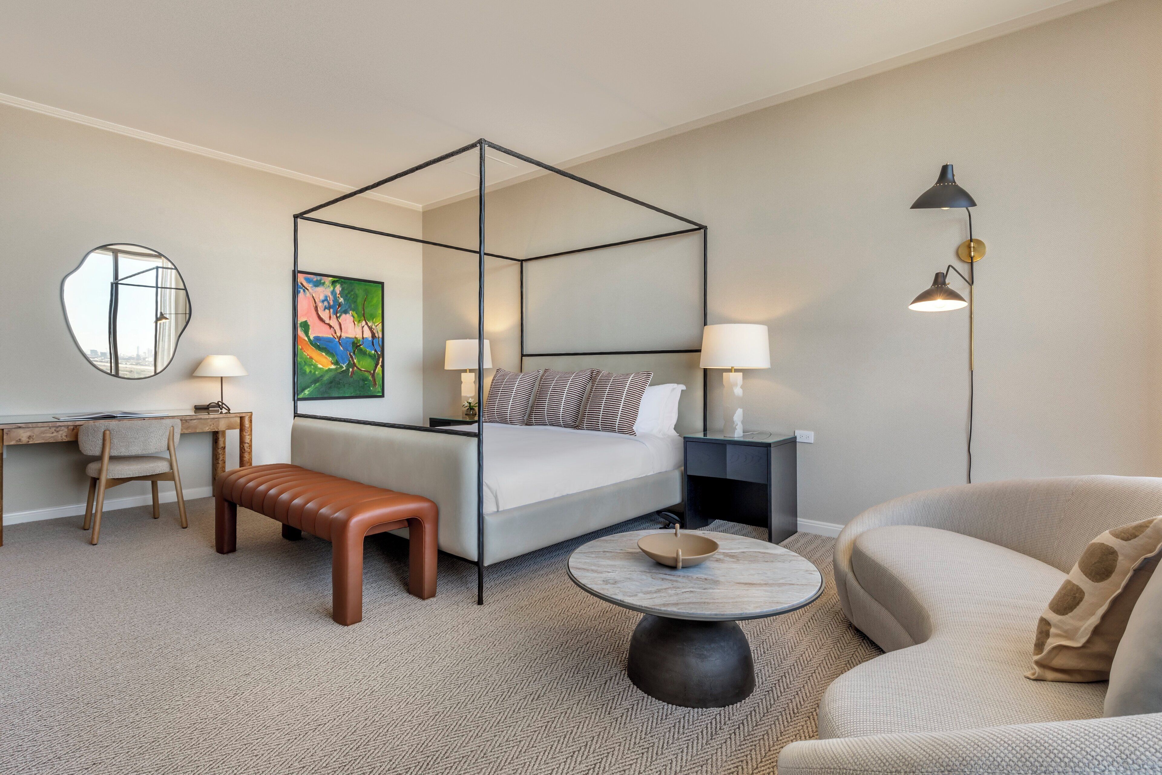 Une chambre d’hôtel moderne avec un lit à baldaquin, un bureau avec une chaise, une table basse ronde, un canapé et un lampadaire.