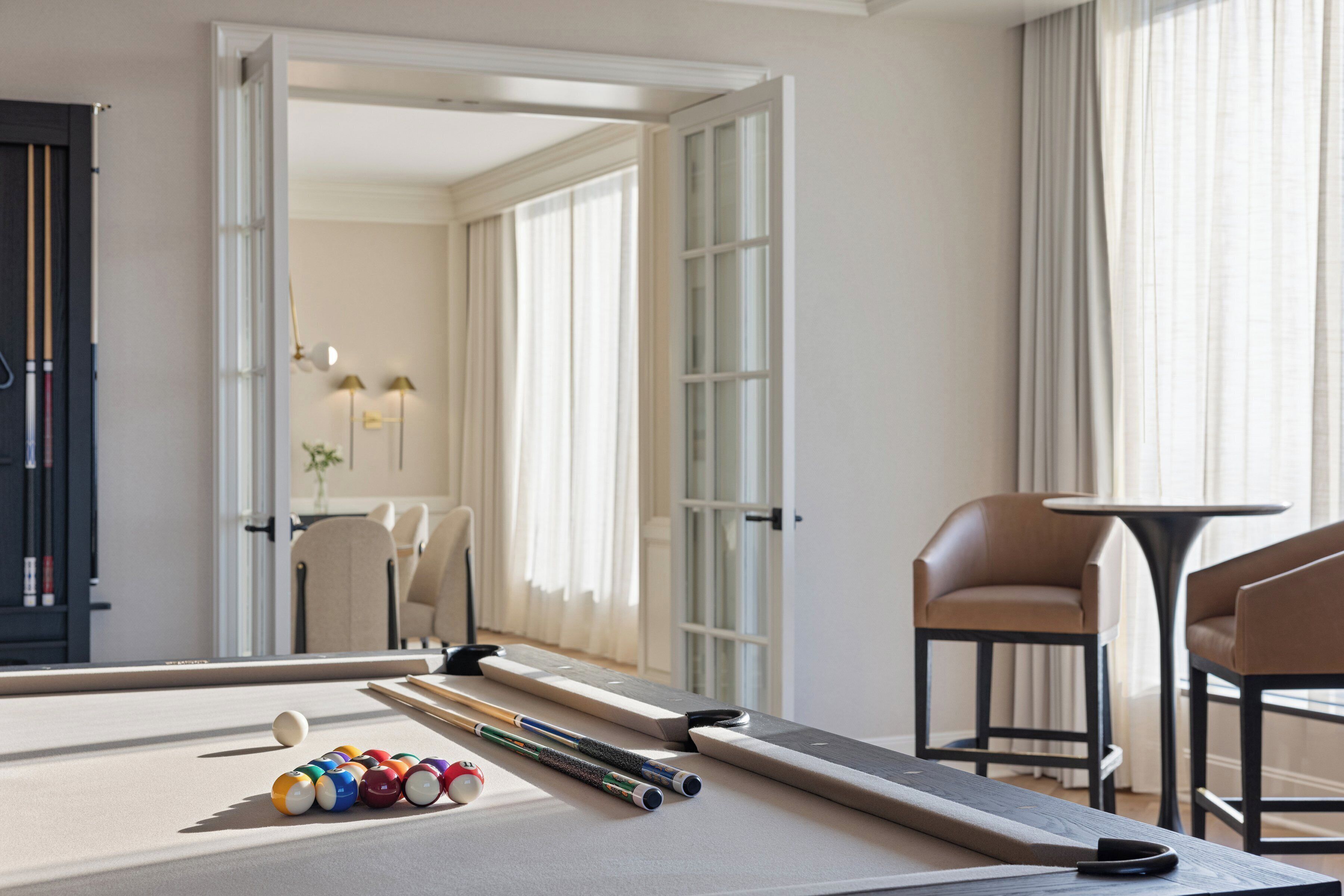 Ein Billardtisch mit Billardkugeln und Spielstangen, ein Sofa und ein Sessel in einem hellen Raum mit großen Fenstern und Vorhängen.