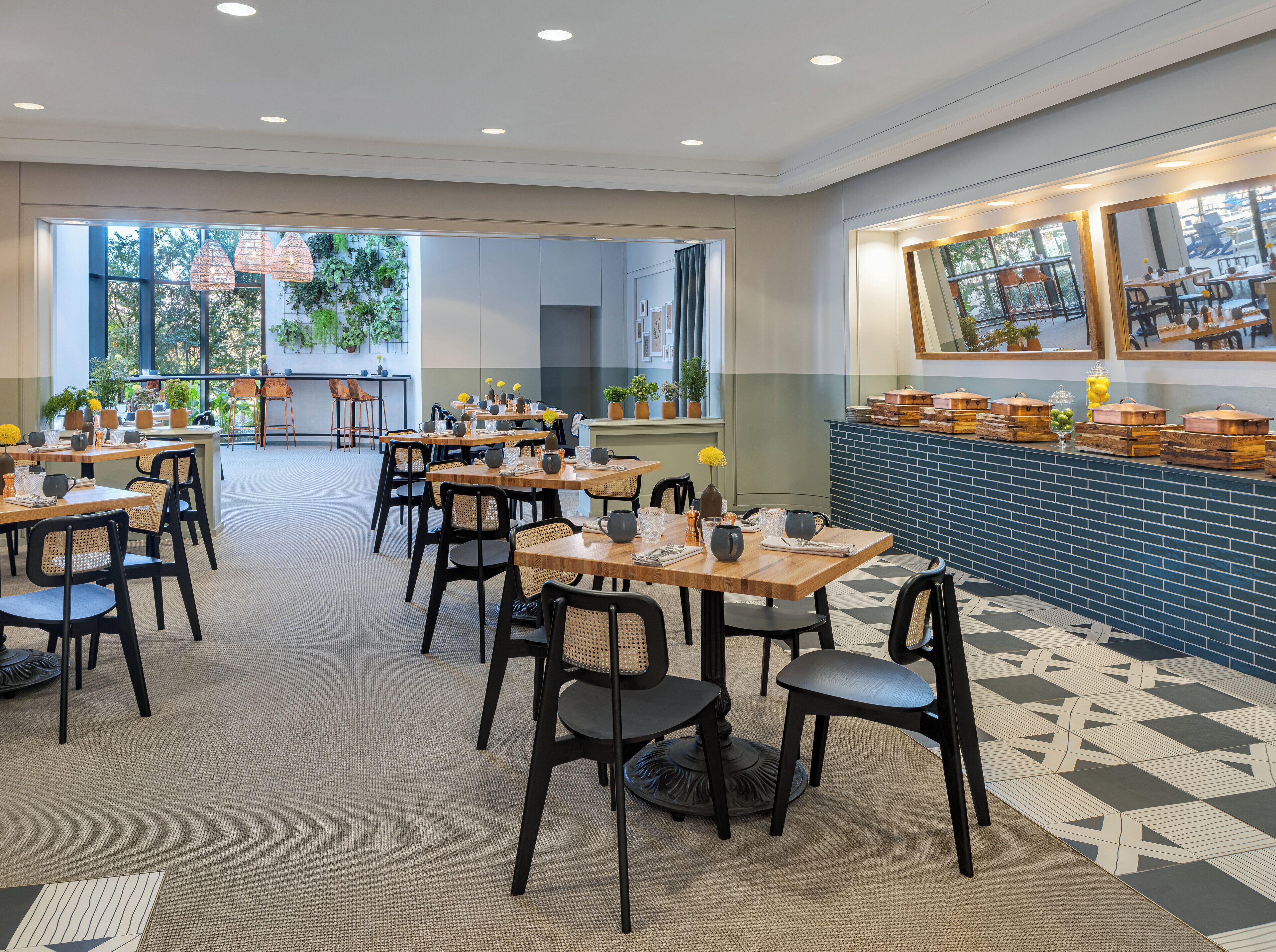 A spacious dining area with multiple tables and chairs, large windows with plants, and a blue-tiled counter with food containers.