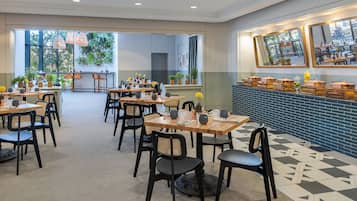 A spacious dining area with multiple tables and chairs, large windows with plants, and a blue-tiled counter with food containers.