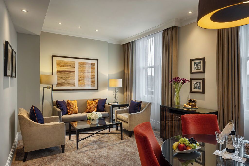A modern hotel room with a sofa set, a dining table with fruit and glasses, a vase with flowers, and a large window with curtains.