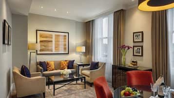 Una habitación de hotel moderna con un conjunto de sofá, una mesa de comedor con fruta y vasos, un jarrón con flores y un gran ventanal con cortinas.