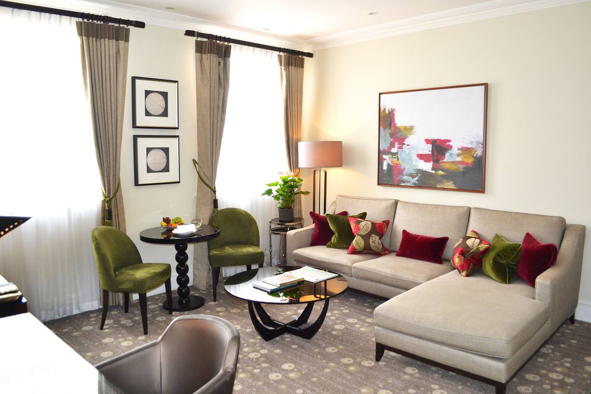 Une chambre d’hôtel moderne, équipée d’un canapé, de deux fauteuils, d’une table basse et d’une table d’appoint avec une lampe. Deux tableaux encadrés ornent le mur, et une grande peinture abstraite est accrochée au-dessus du canapé.
