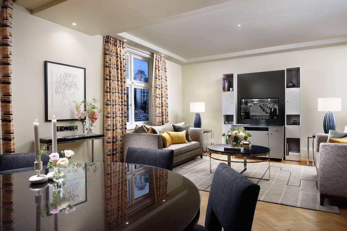 Un salon moderne avec un canapé, une table basse et un téléviseur. On y trouve une table à manger décorée de bougies et de fleurs. La pièce est dotée d’une grande fenêtre avec des rideaux.
