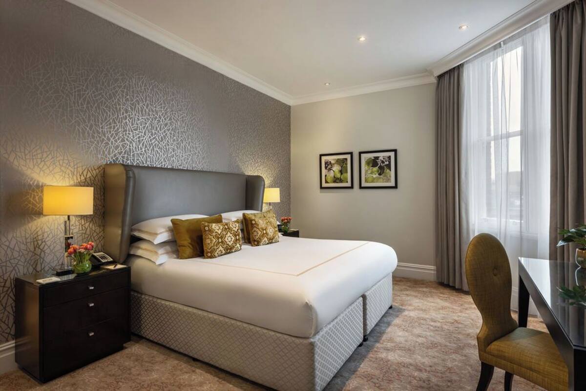 Une chambre d’hôtel avec un grand lit, une table de chevet avec une lampe, deux tableaux encadrés au mur et un coin repas avec une chaise.