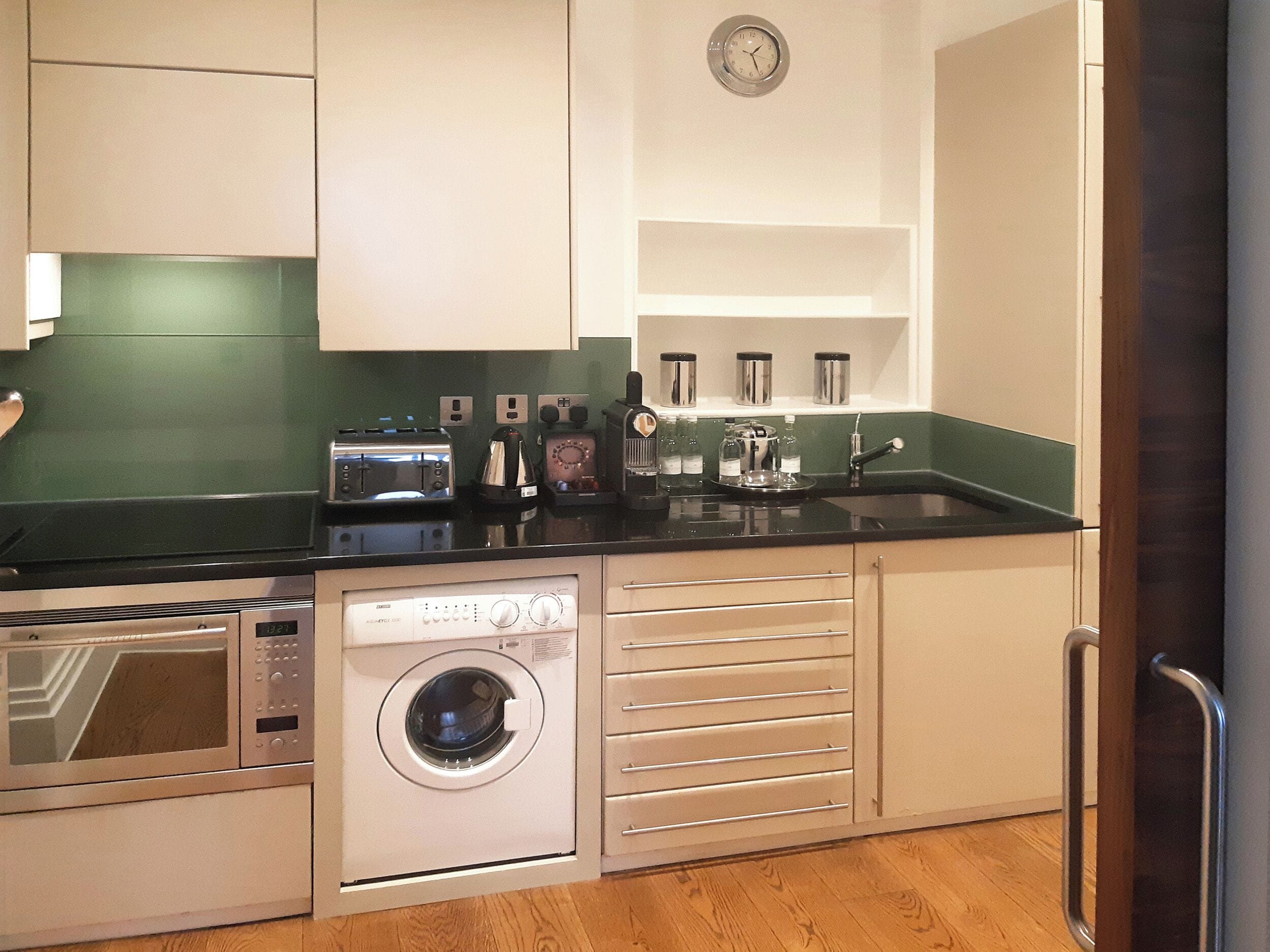A compact kitchen with a washing machine, a toaster, and a clock on the wall.