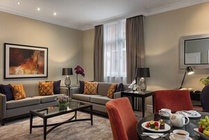 A modern living room with a sofa, coffee table, and dining area. There is a painting on the wall, a window with curtains, and a lamp on a side table.