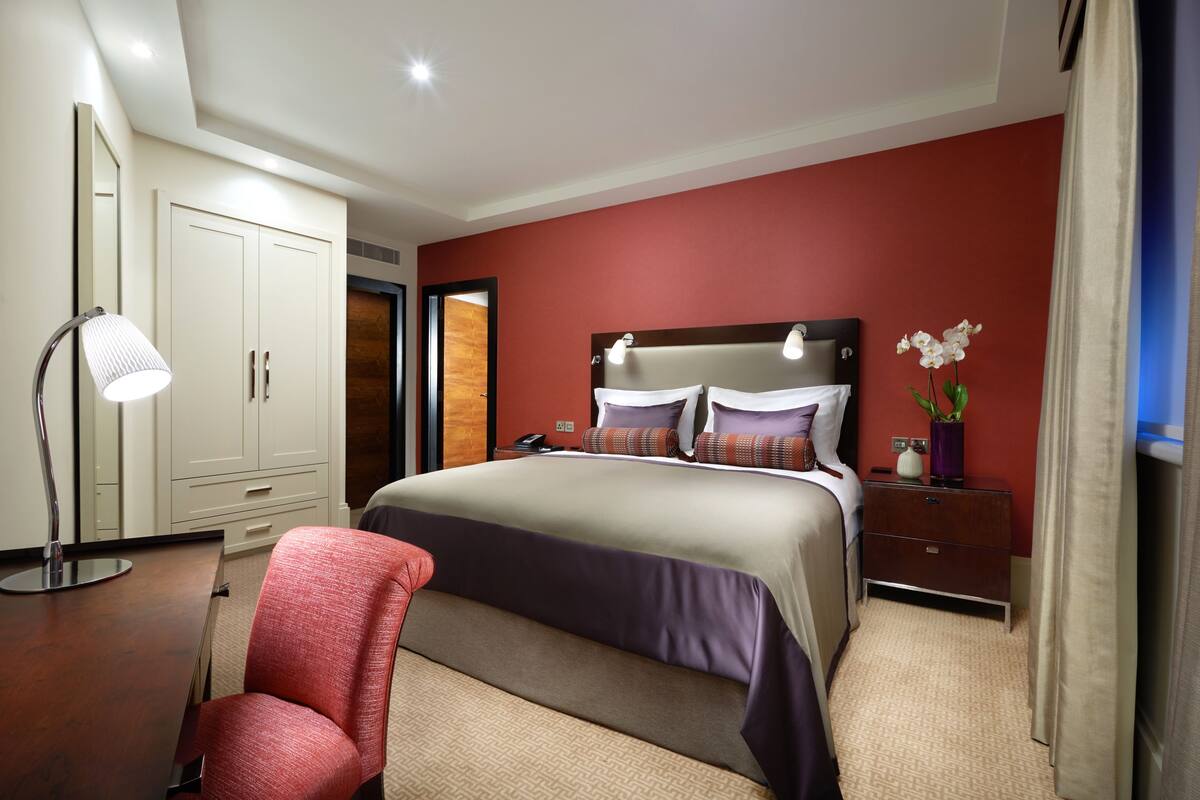 Une chambre d’hôtel avec un grand lit, un bureau avec une lampe, une armoire et une table de chevet agrémentée d’un vase de fleurs.