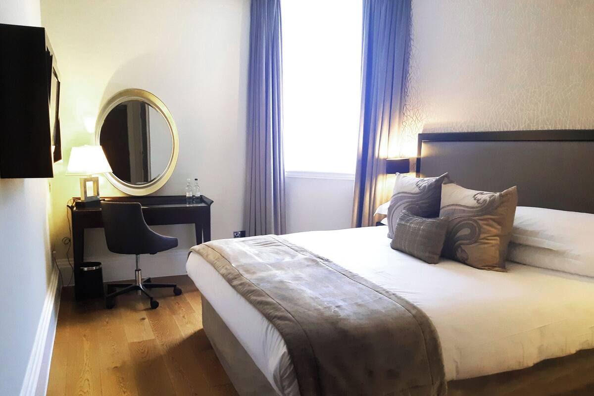 Une chambre d’hôtel avec un lit, un bureau et une chaise, un miroir et une fenêtre avec des rideaux.