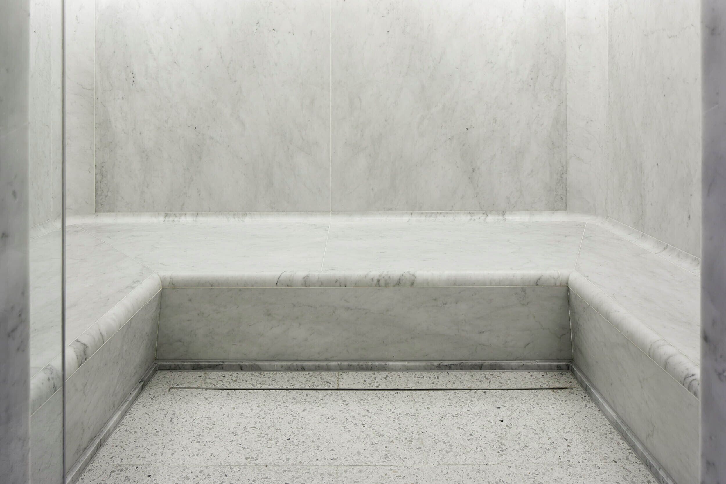 Un baño con paredes y suelo de mármol blanco, un rincón ducha y un escalón pequeño que conduce a la ducha.