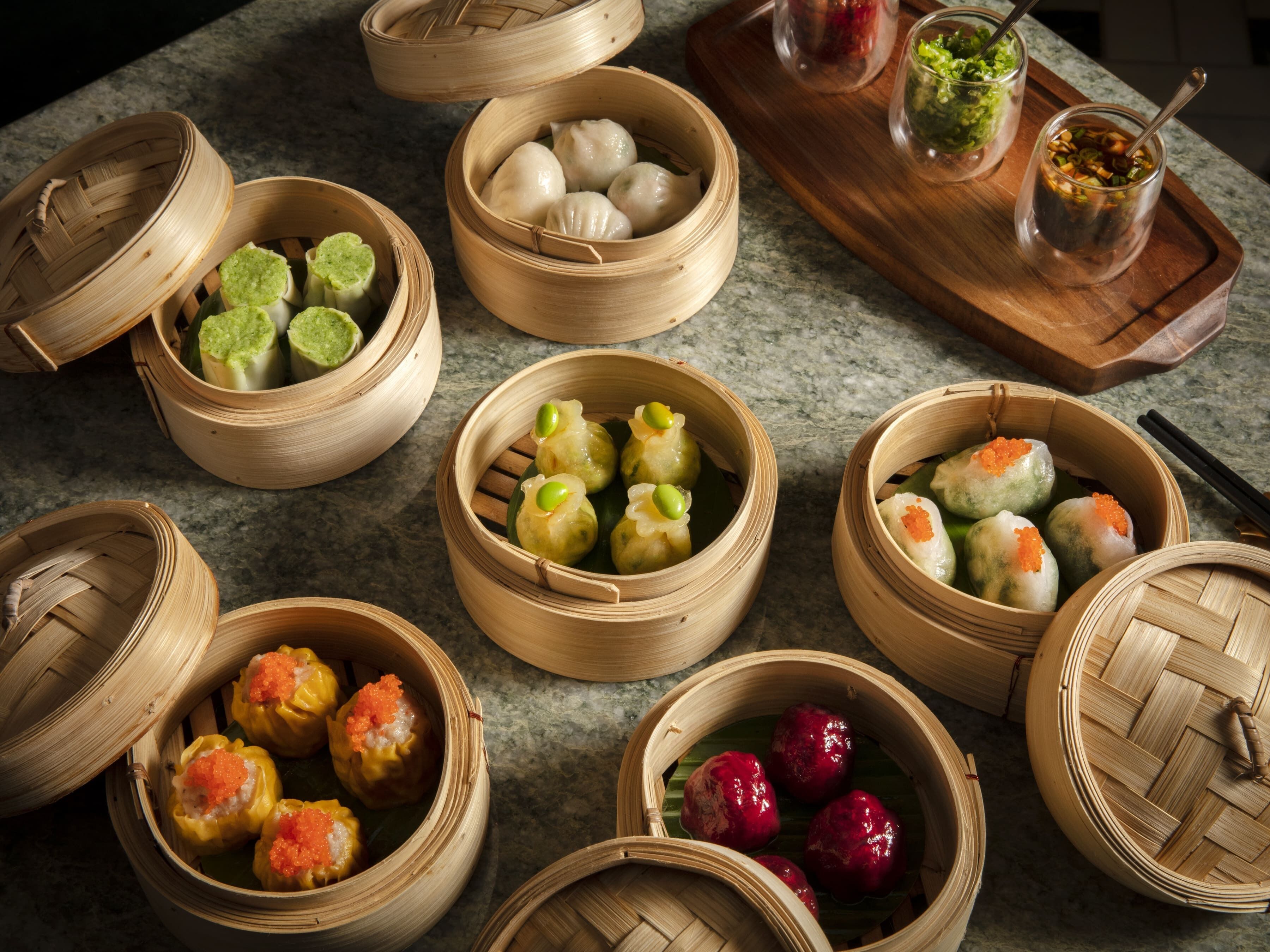 Une variété de plats de dim sum, présentés dans des paniers à vapeur en bambou, notamment des raviolis verts, jaunes et rouges, disposés sur un comptoir.