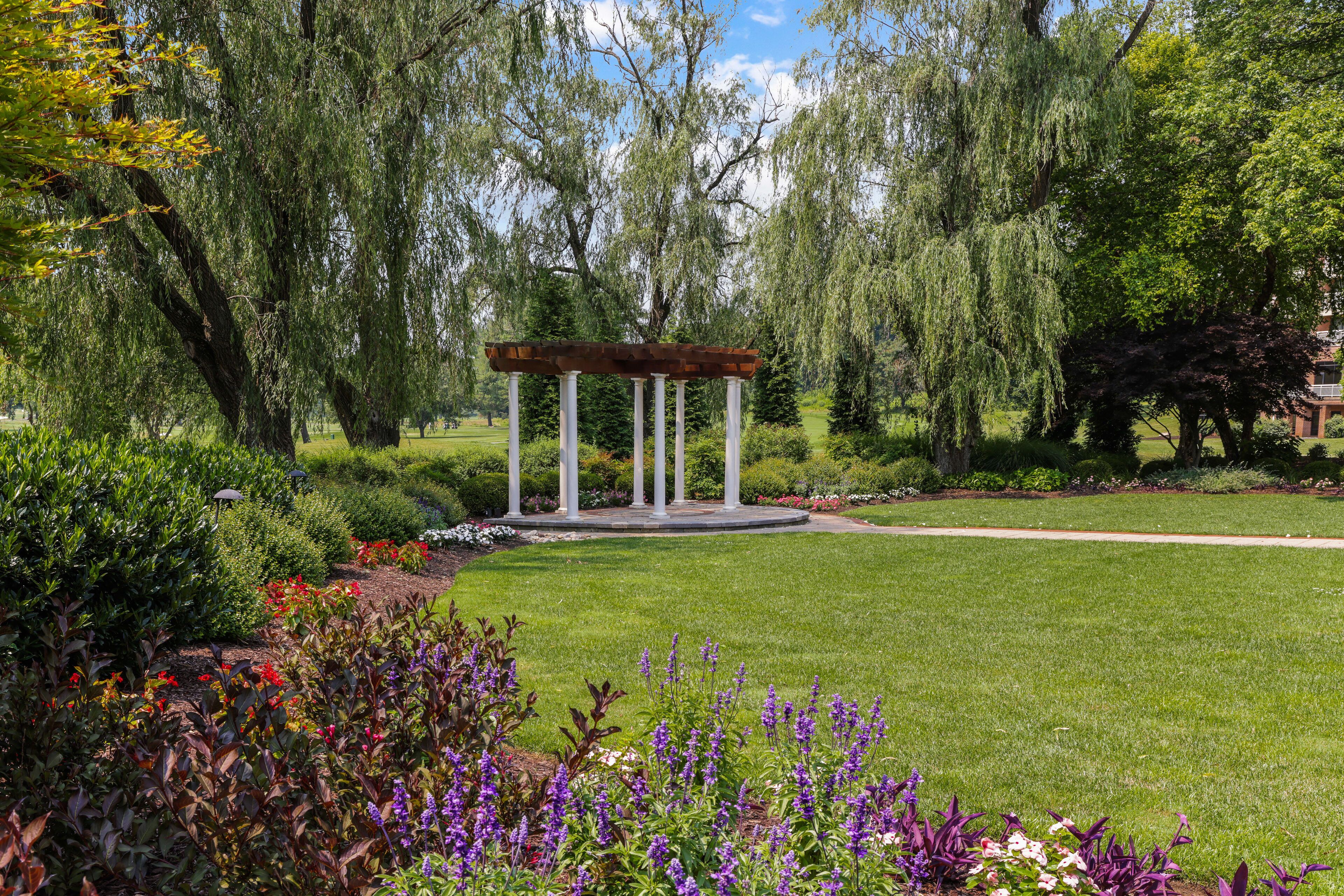 Un quiosco con base circular y columnas blancas, rodeado de abundante follaje y plantas en flor.