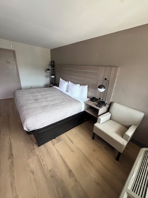 A hotel room with a large bed, a bedside table with a lamp, a chair, and a radiator.