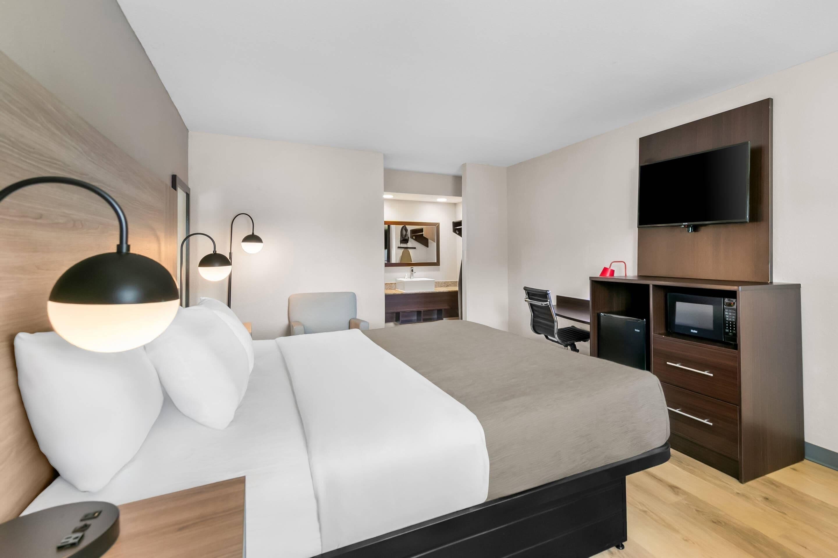 Moderni hotellihuone, jossa on sänky, yöpöytävalaisimet, televisio, minibaari ja työpöytä.