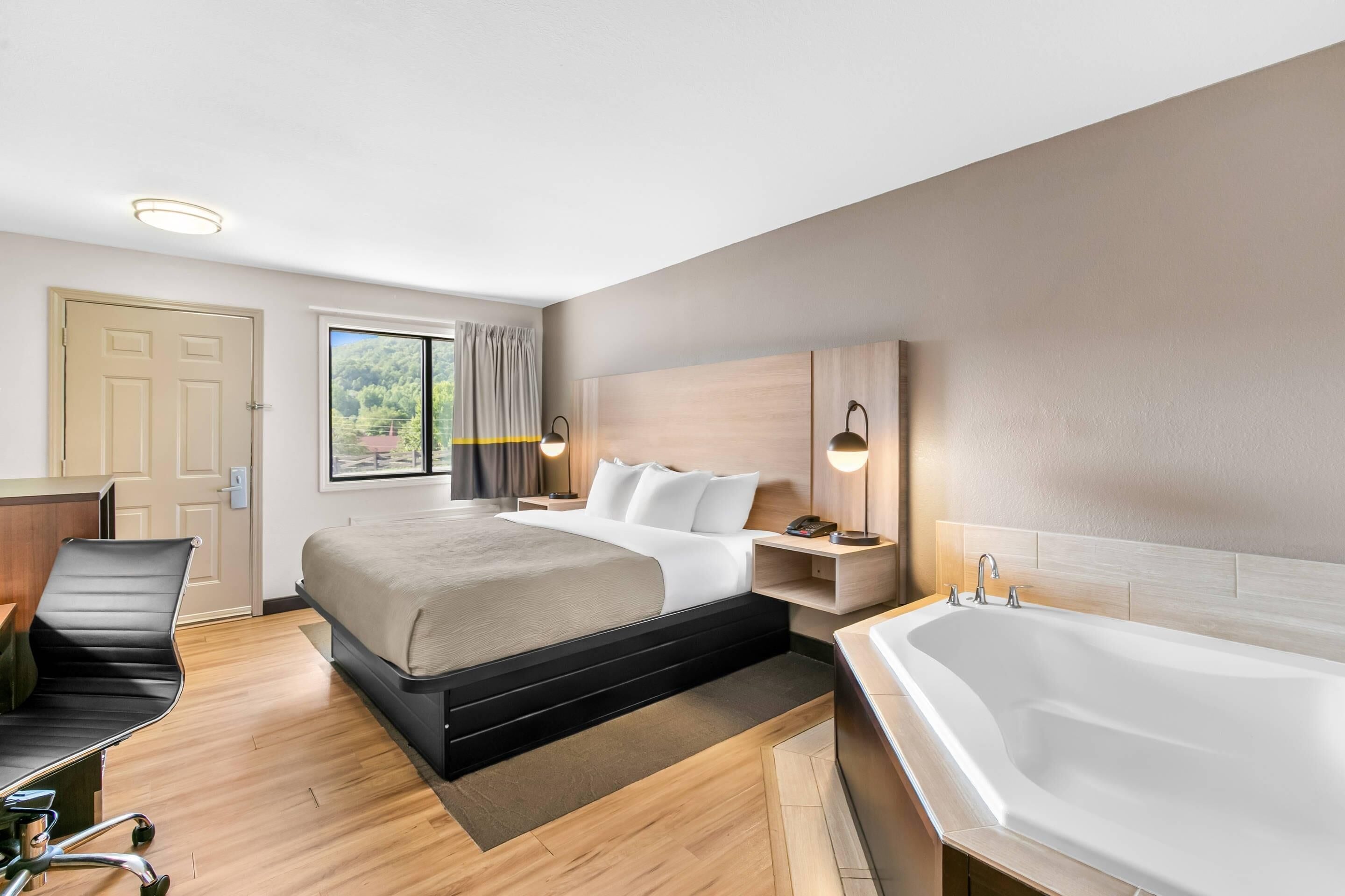 A modern hotel room with a bed, bedside tables, a desk, a chair, a large window, and a bathtub.