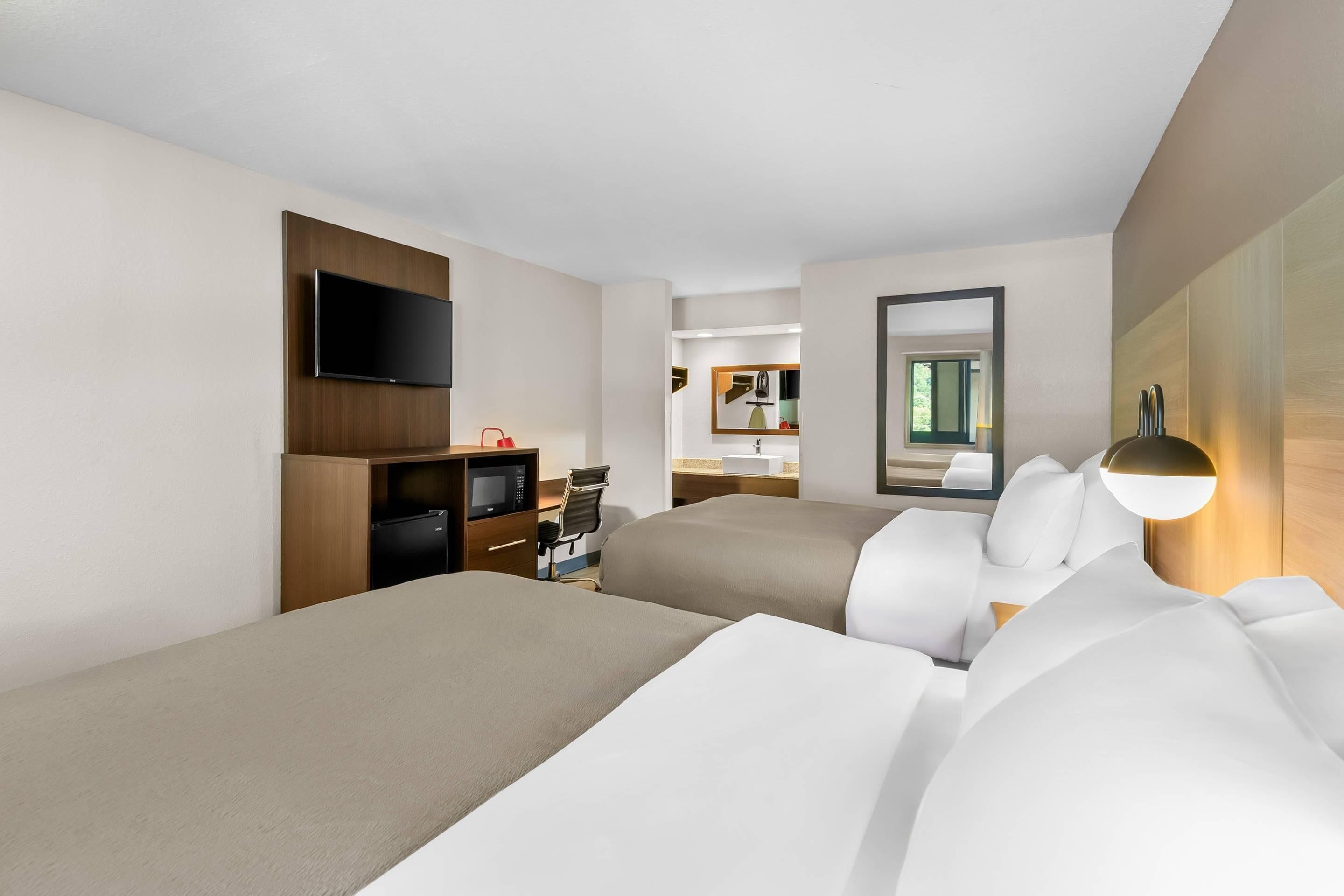 A hotel room with two beds, a TV, a desk with a microwave, and a bathroom visible through an open door.