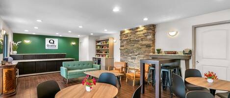A hotel lobby with a reception desk, a green wall with a 'Quality by Choice Hotels' sign, a fireplace, and a dining area with tables and chairs.