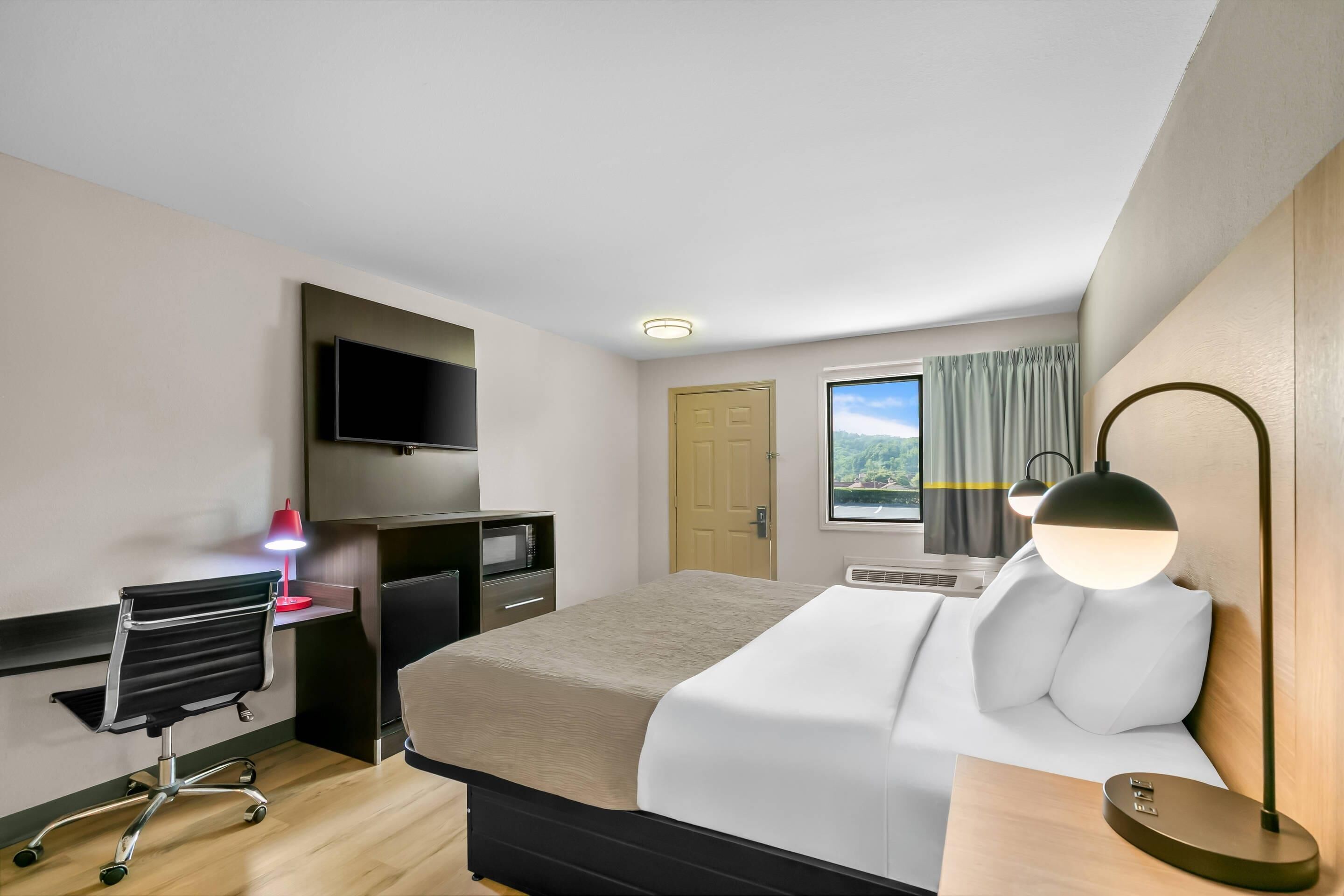 A hotel room with a bed, a desk with a chair, a television, a window with curtains, and a lamp.