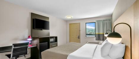 A hotel room with a bed, a desk with a chair, a television, a window with curtains, and a lamp.