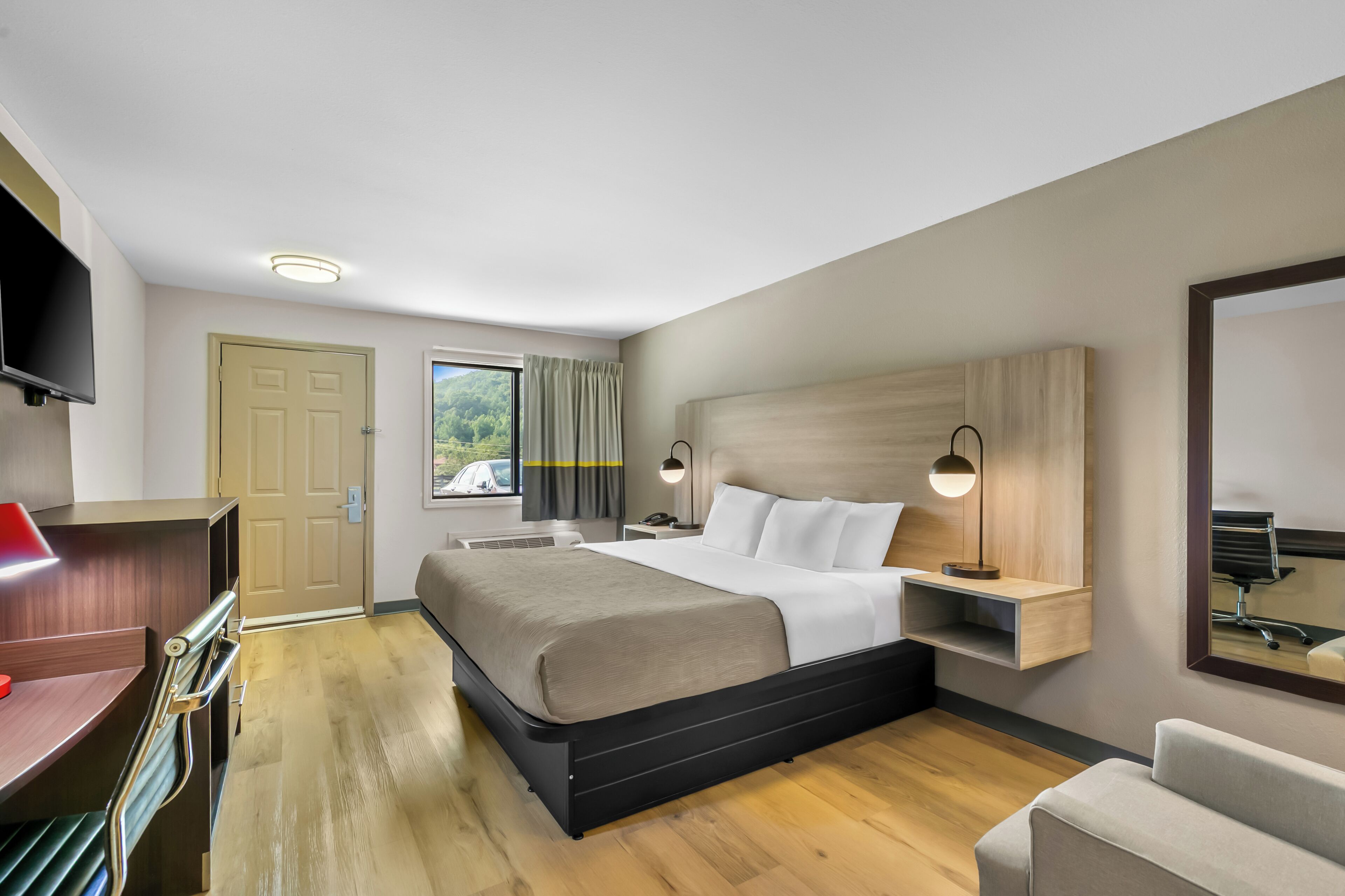 A hotel room with a bed, bedside table, lamp, TV, desk, chair, and a window with curtains.
