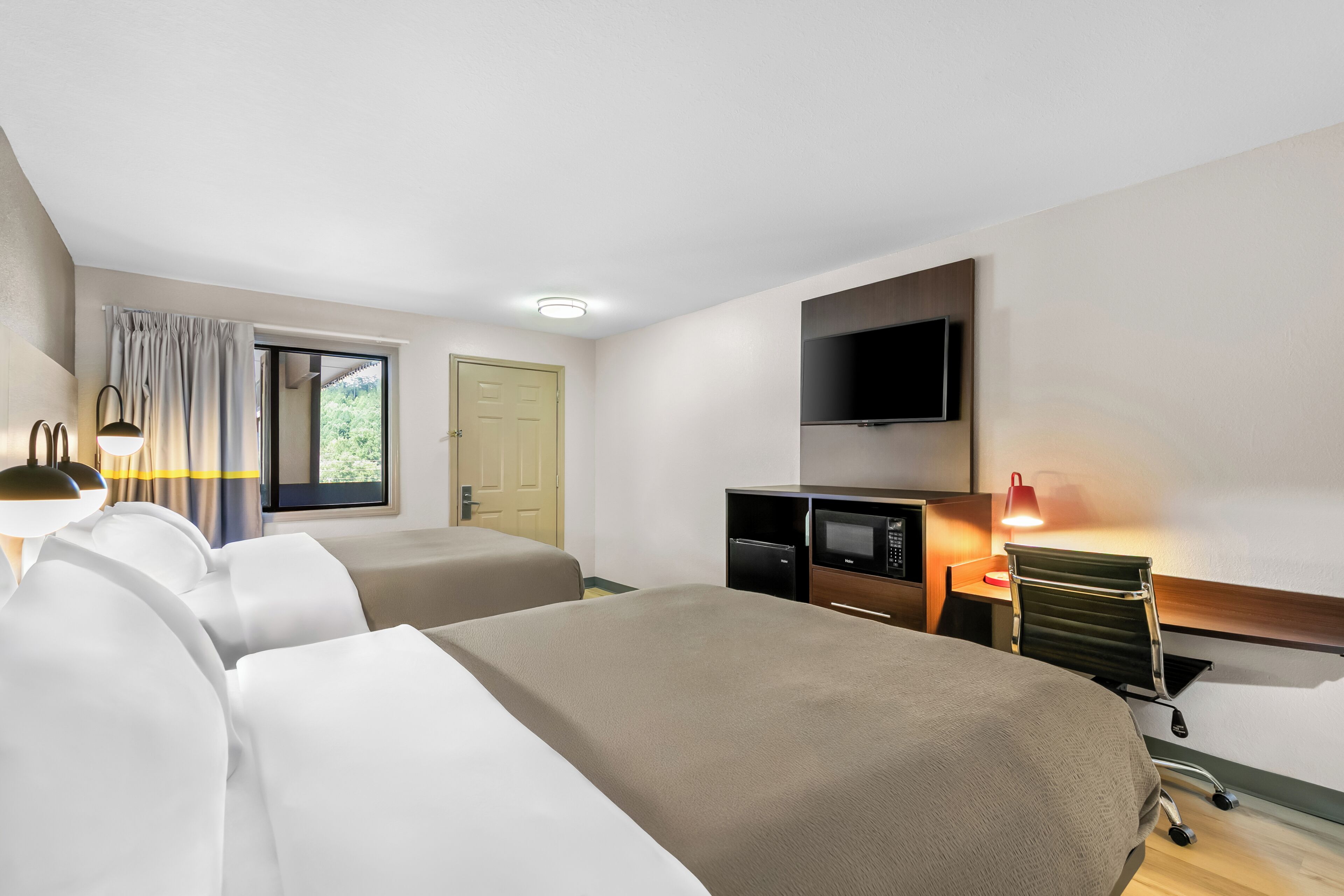 A hotel room with two beds, a flat-screen TV, a microwave, and a desk with a lamp.