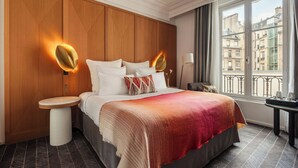 Une chambre d’hôtel avec une tête de lit en bois, un lit recouvert de draps blancs, un plaid rouge aux nuances dégradées, une table de chevet avec une lampe, et une vue sur les bâtiments par la fenêtre.