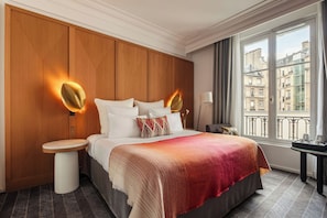Ein Hotelzimmer mit massivem Holzkopfende, einem Bett mit weißer Bettwäsche, einer roten Farbverlaufdecke, einem Nachttisch mit Lampe und Blick auf Gebäude durch das Fenster.