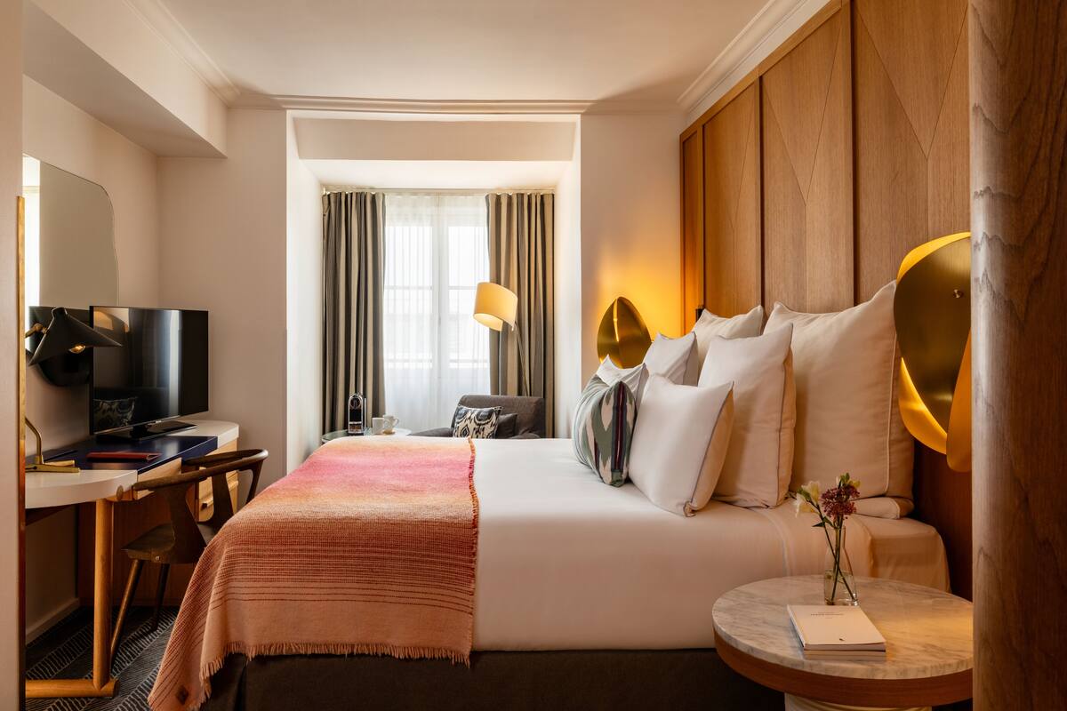 A hotel room with a bed, a desk with a TV, a lamp, and a bedside table with a book and flowers.