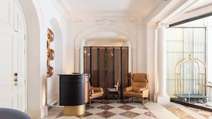 Un hall d’entrée avec un sol à carreaux, un meuble noir et doré, deux fauteuils en cuir, une cloison en verre et une fenêtre ornée d’un cadre doré.